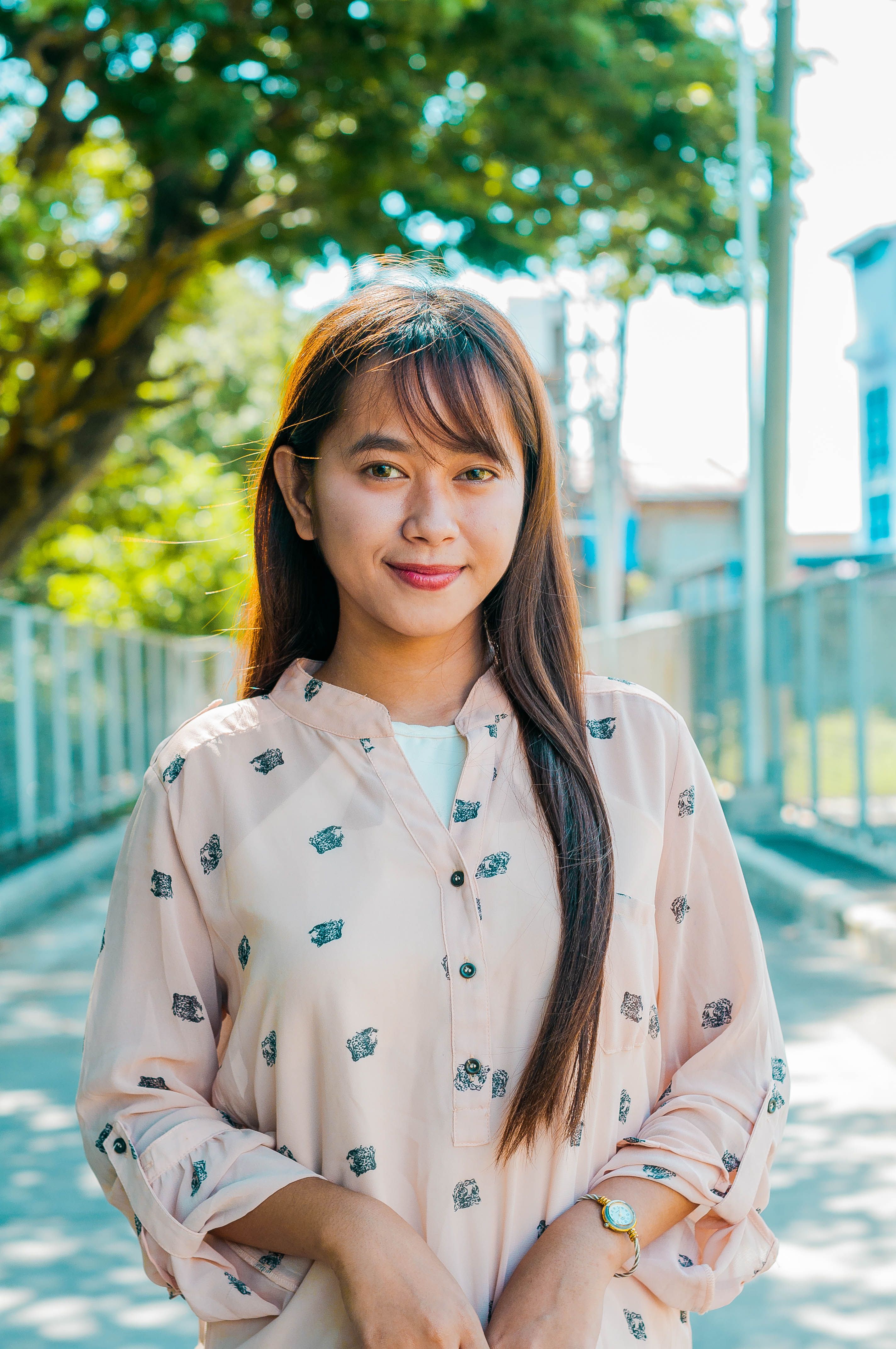 Charming young Asian female in elegant outfit looking at camera while standing on pathway with fences and trees on street