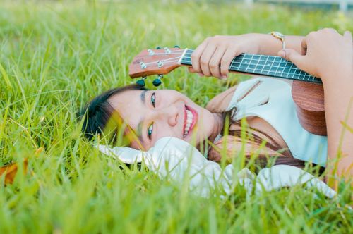 Free Delighted Asian female musician with ukulele smiling and looking at camera while lying on green grassy lawn on summer day Stock Photo