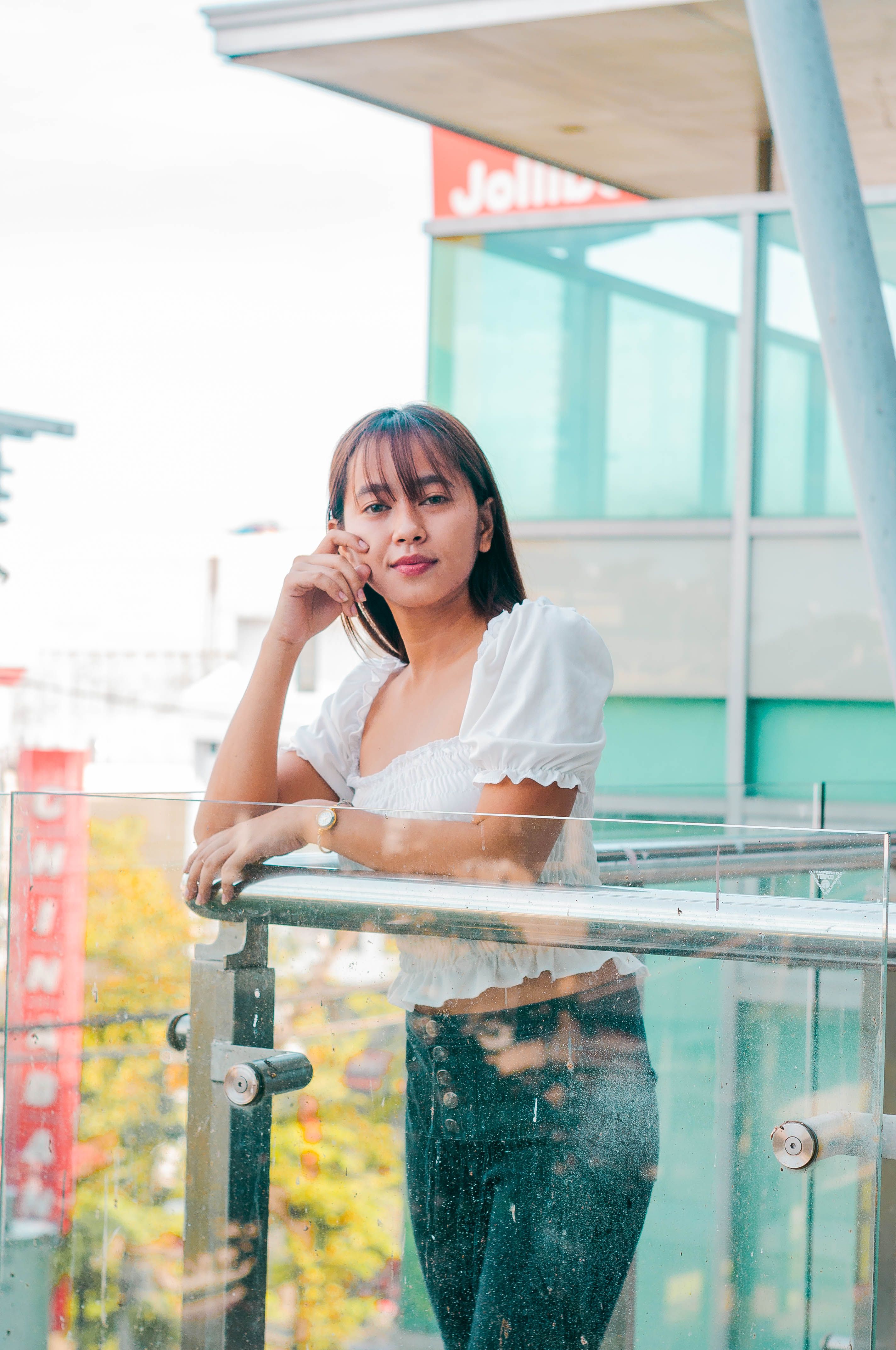 Young Asian female in trendy outfit leaning on railing and looking at camera while standing near modern building in city