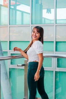 Joyful Asian female in stylish outfit looking at camera while standing at metal railing near contemporary house with glass wall