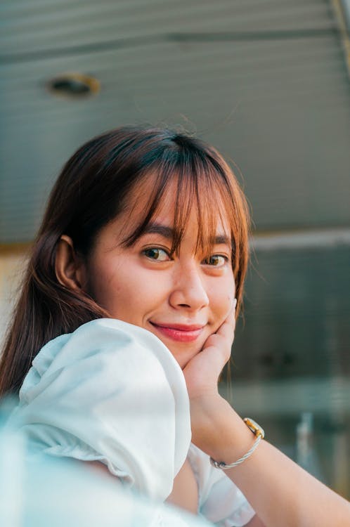 Cheerful Asian female with long brown hair touching cheek and looking at camera while standing at blurred railing near building