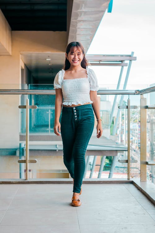 Full body of smiling Asian female in stylish white top and jeans looking at camera while strolling on glass balcony
