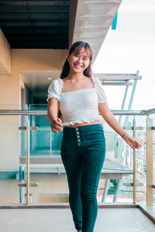 Free Young Asian female with long hair in trendy outfit looking at camera with smile while walking on fenced balcony with metal railing Stock Photo