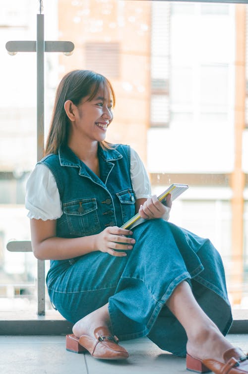 Free Cheerful young Asian female with notepad in hands wearing denim outfit sitting on floor near big window of modern building Stock Photo