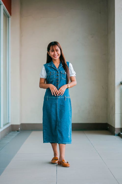 Full body of positive Asian female with long hair wearing denim dress looking at camera while standing on terrace of building