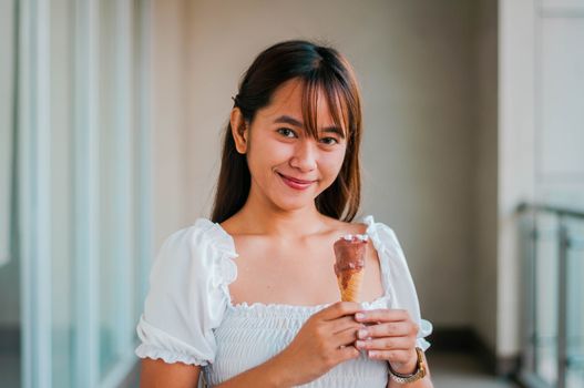 Cheerful Asian female in white outfit with sweet chocolate ice cream cone in hand looking at camera while standing on balcony
