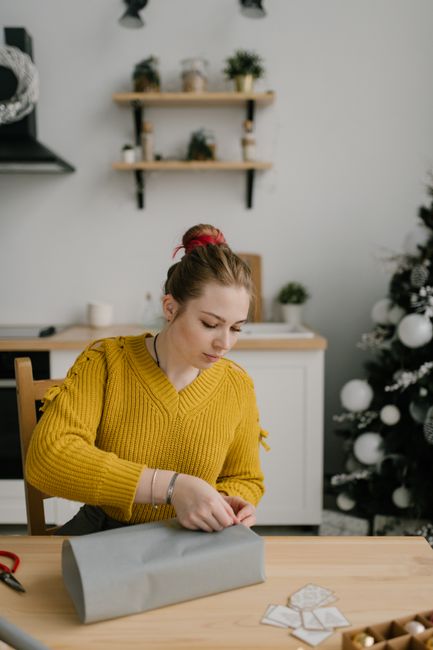 A woman in a cozy sweater wraps gifts at a table with a Christmas tree in the background.