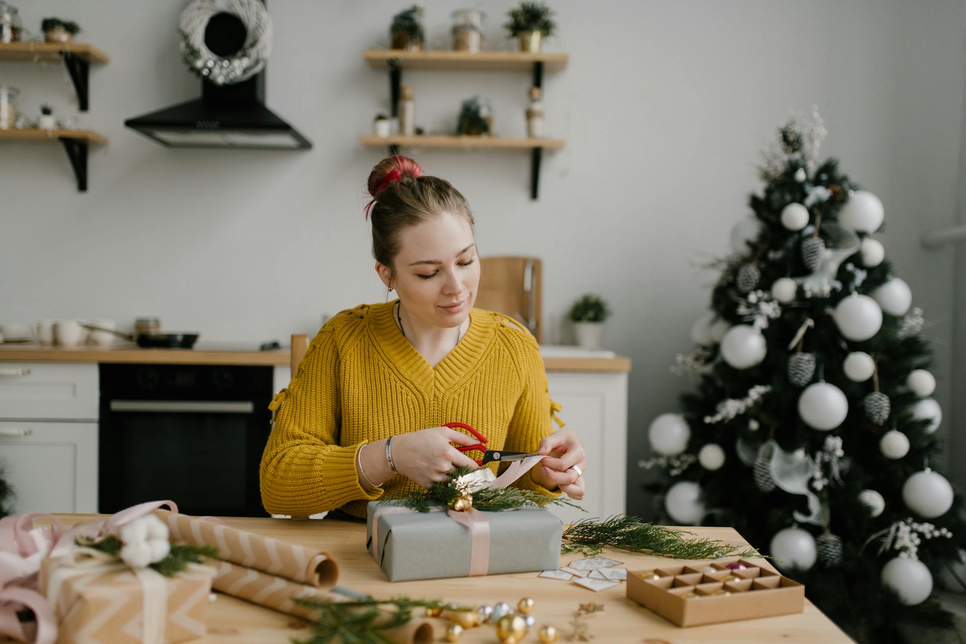 A woman in a yellow sweater wrapping Christmas presents in a modern kitchen with a decorated tree.