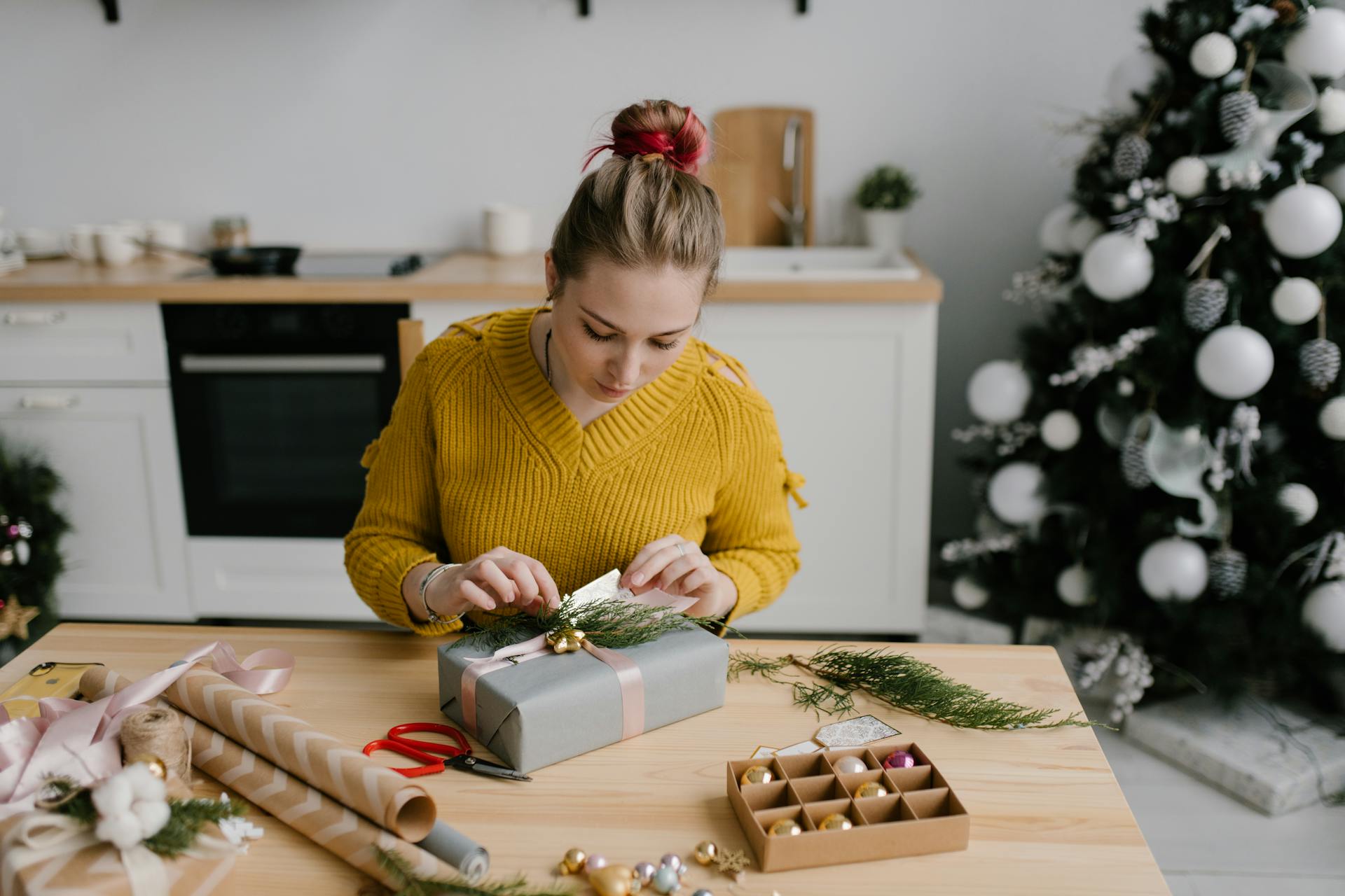 A woman in a yellow sweater wraps a Christmas gift in a festive kitchen setting.