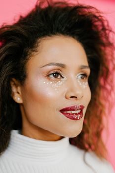 Vibrant portrait of an African American woman with artistic makeup and glittery lips on a pink backdrop.