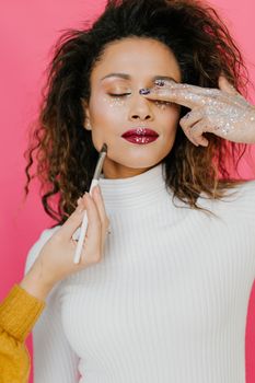 Close-up portrait of a woman applying glitter makeup in a white turtleneck.