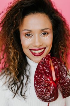 Portrait of a smiling woman in a white turtleneck holding a red anthurium against a pink background.