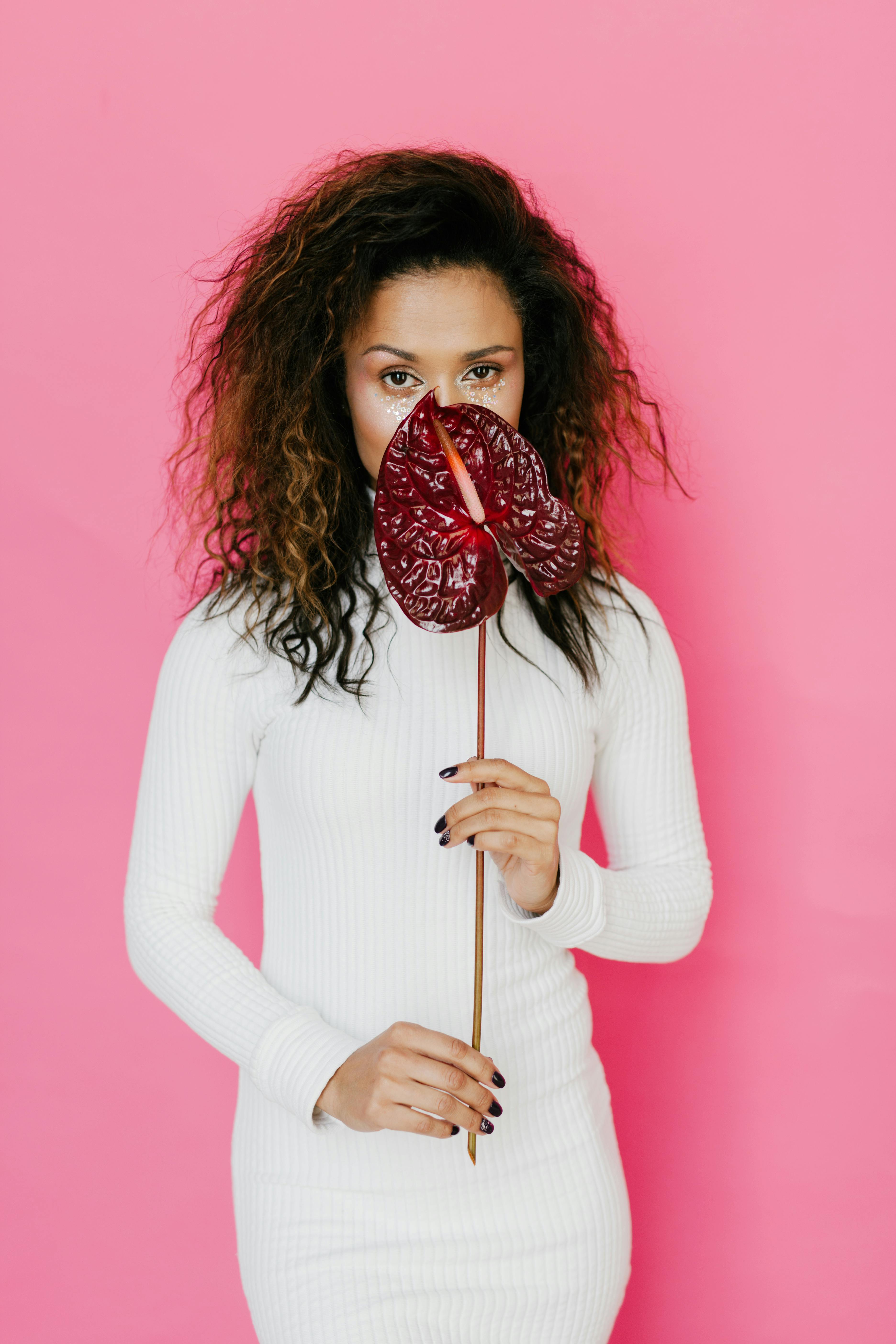 一位时尚女子身着白色连衣裙，手持红色红掌，背景是充满活力的粉红色