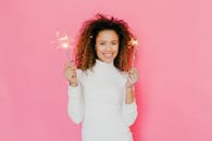 一位快乐的女子身穿白色高领毛衣，在粉色背景下举着烟花庆祝