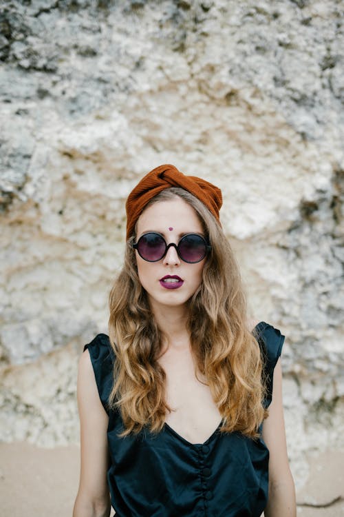 Portrait of serious lady in sunglasses looking at camera and standing against white stone wall in daytime