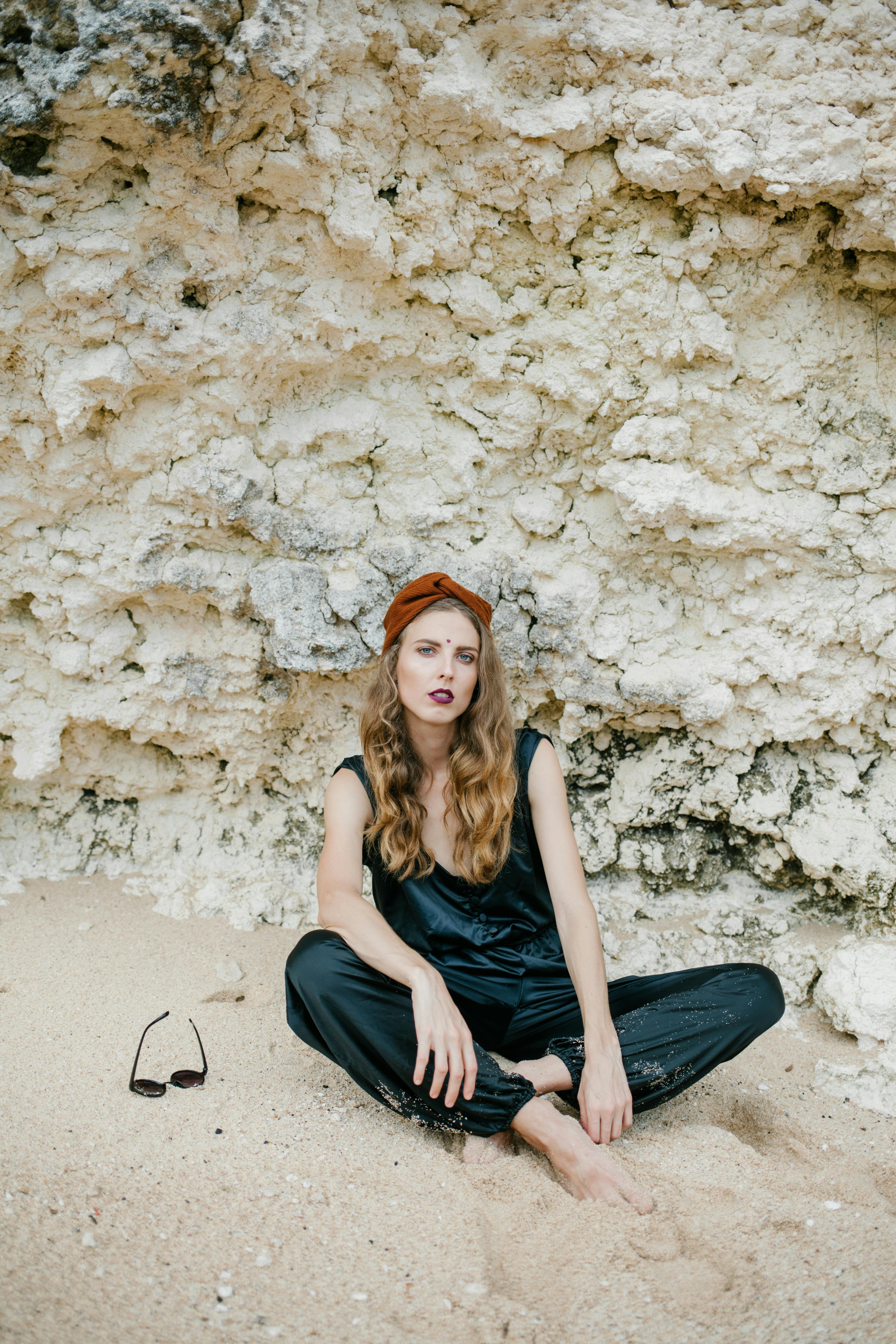 Full body of barefoot female with curly hair and toes in sand sitting against mineral formation