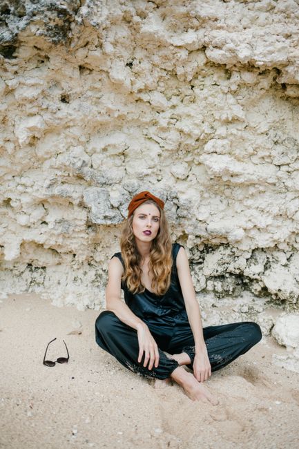 Full body of barefoot female with curly hair and toes in sand sitting against mineral formation