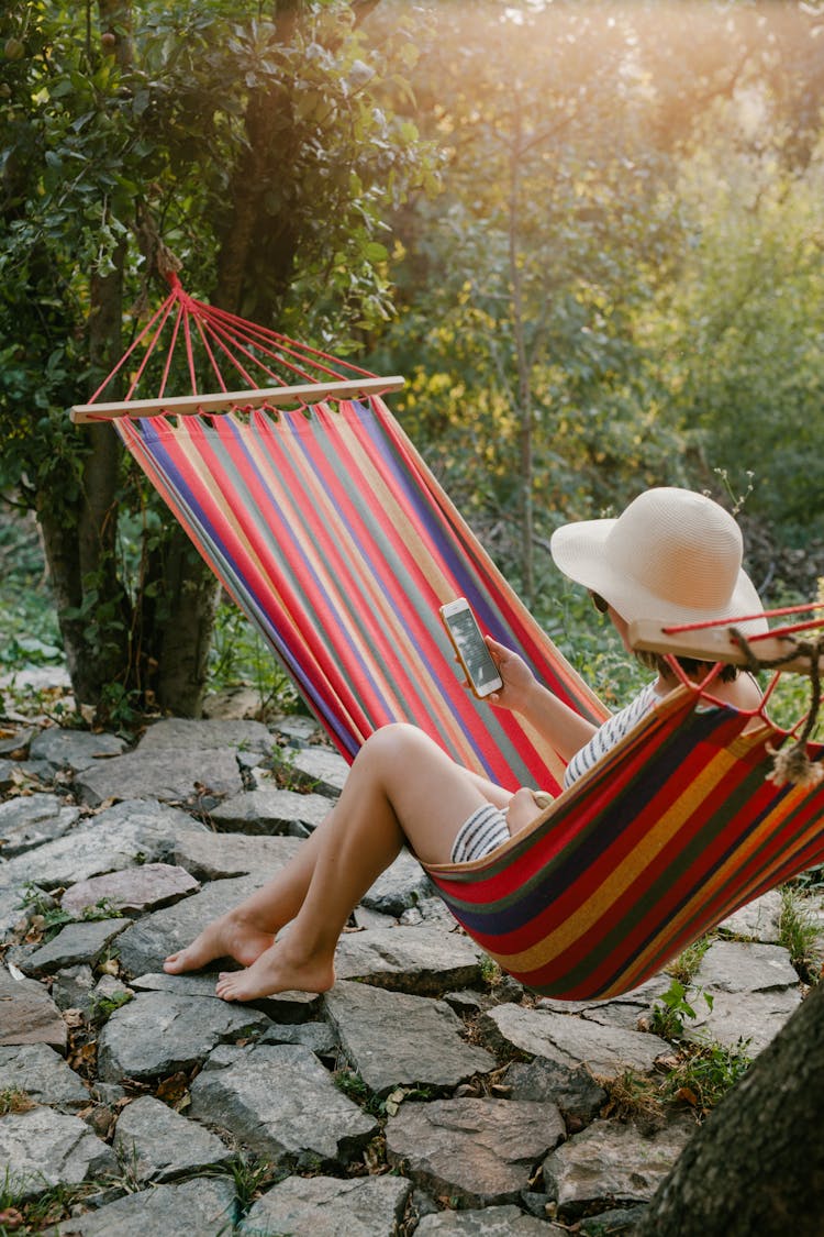 Woman With Smartphone Sitting On Hammock In Green Garden