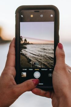 Hands holding smartphone capturing scenic sunset beach view, showcasing mobile photography.