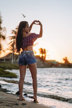 Woman on the beach making a heart with her hands at sunset, capturing the essence of a tropical summer vacation.