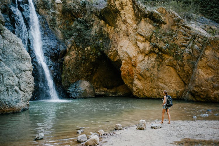 Anonymous Backpacker Contemplating Fast Waterfall In Mountains Near Pond