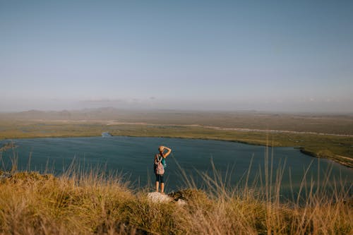 Back view of anonymous female backpacker admiring rippled river from mountain with faded grass on foggy day