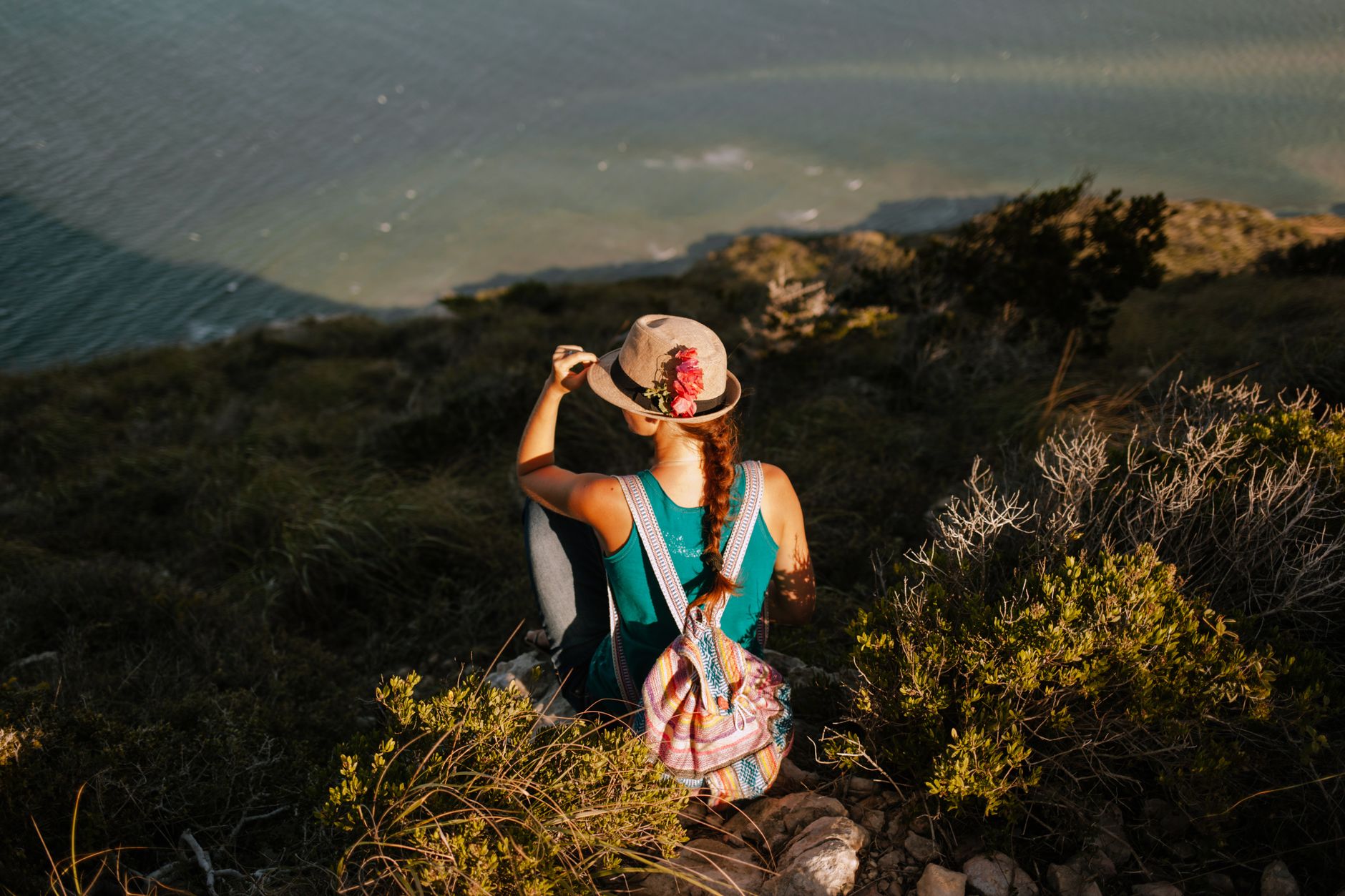 From above back view of unrecognizable traveler with backpack touching straw hat while admiring river from grass mountain