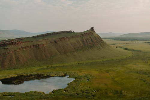 Scenic view of mounts near grass meadow with small pond reflecting foggy sky in daylight