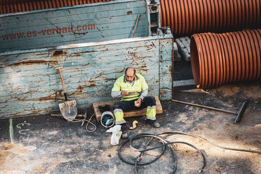 A construction worker sitting on a site in Madrid, enjoying a break amid tools and pipes.
