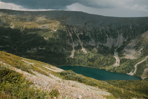 Picturesque rocky mountains covered with grass and bushes near clean blue lake under cloudy gloomy sky