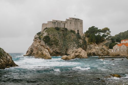 Free Ancient gray castle located on rough rocky stony cliff covered with green bushes and trees washed by wavy foamy ocean Stock Photo