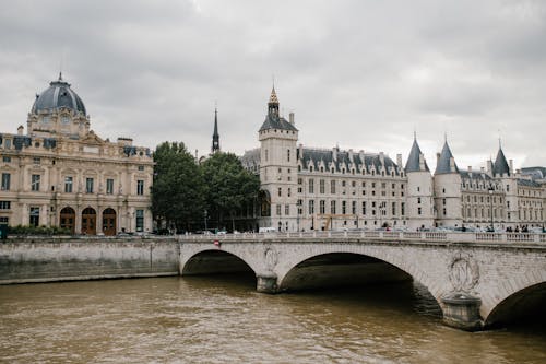 Aged stone bridge crossing river with architectural monument Conciergerie castle located in Paris under cloudy sky