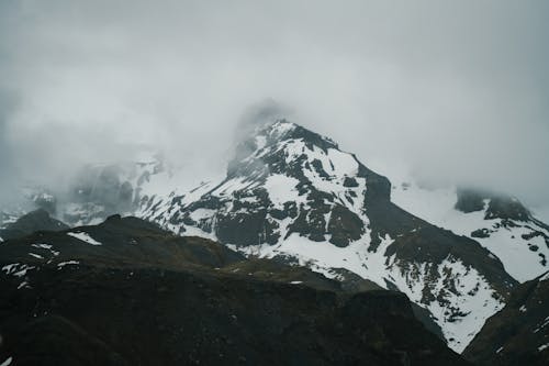 Scenic view of rough rocky mountains with slopes covered with snow and peaks in dense fog on overcast weather