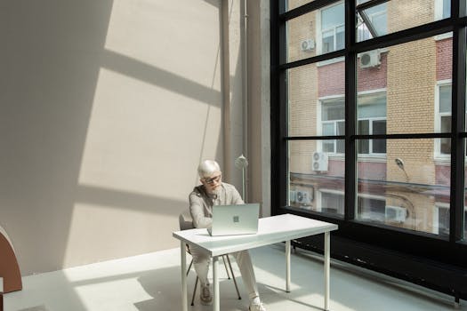 A focused man works on his laptop in a modern, sunlit office space with large windows.