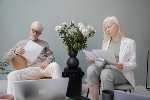 Busy woman and man with white hair sitting against wall near vase with flowers while reading papers in office during work