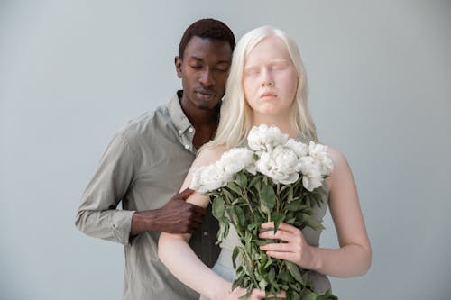 Calm woman with pale skin and bouquet of flowers standing near African American male with closed eyes on gray background
