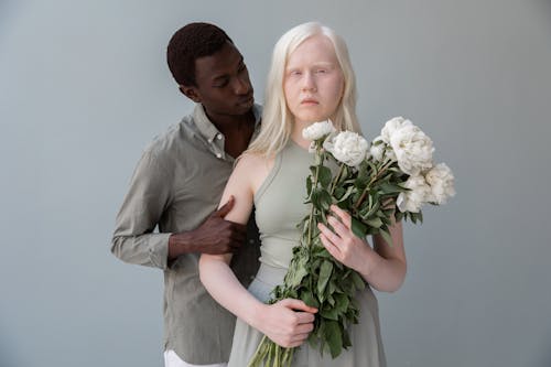 African American male touching arm of woman with pale skin while standing on gray background with bouquet of flowers and looking at camera