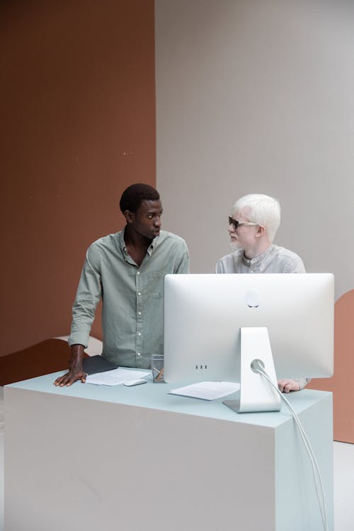 Black man with albino colleague standing near computer and looking at each other while having conversation about work in creative office