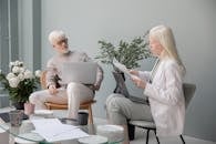 Focused woman with documents sitting against wall in office with table and flowers near man with white beard and laptop while working