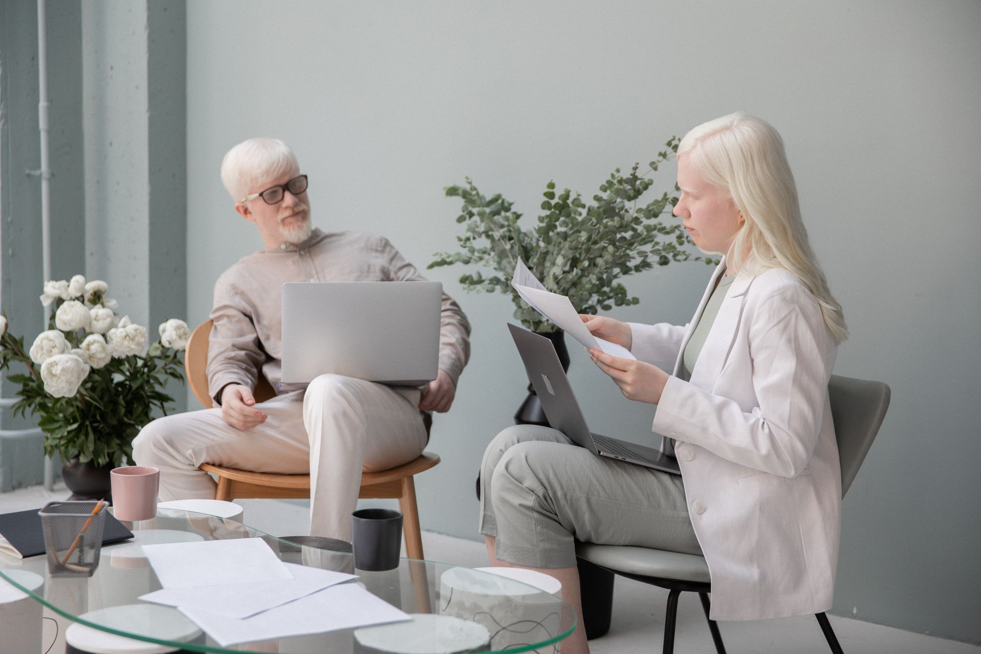 Focused woman with documents sitting against wall in office with table and flowers near man with white beard and laptop while working