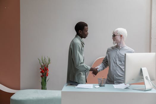 Side view of African American man shaking hand of albino male coworker while standing near table with computer in modern office