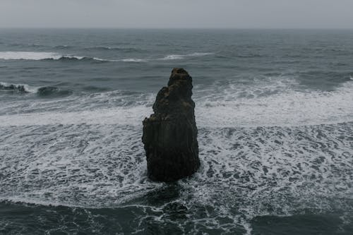 Severe rough rock formation located in shallow sea washed by foamy waves under gloomy sky