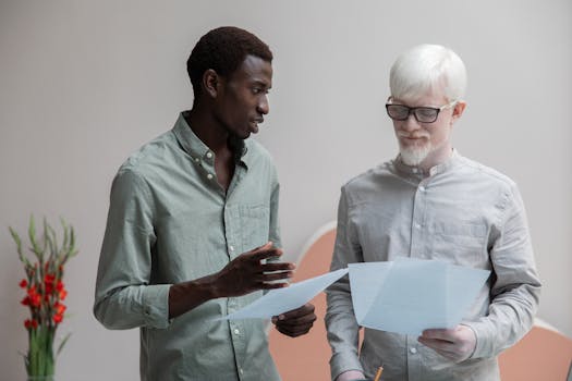 Concentrated African American man and albino coworker in eyeglasses working with papers while discussing work in contemporary office with flower