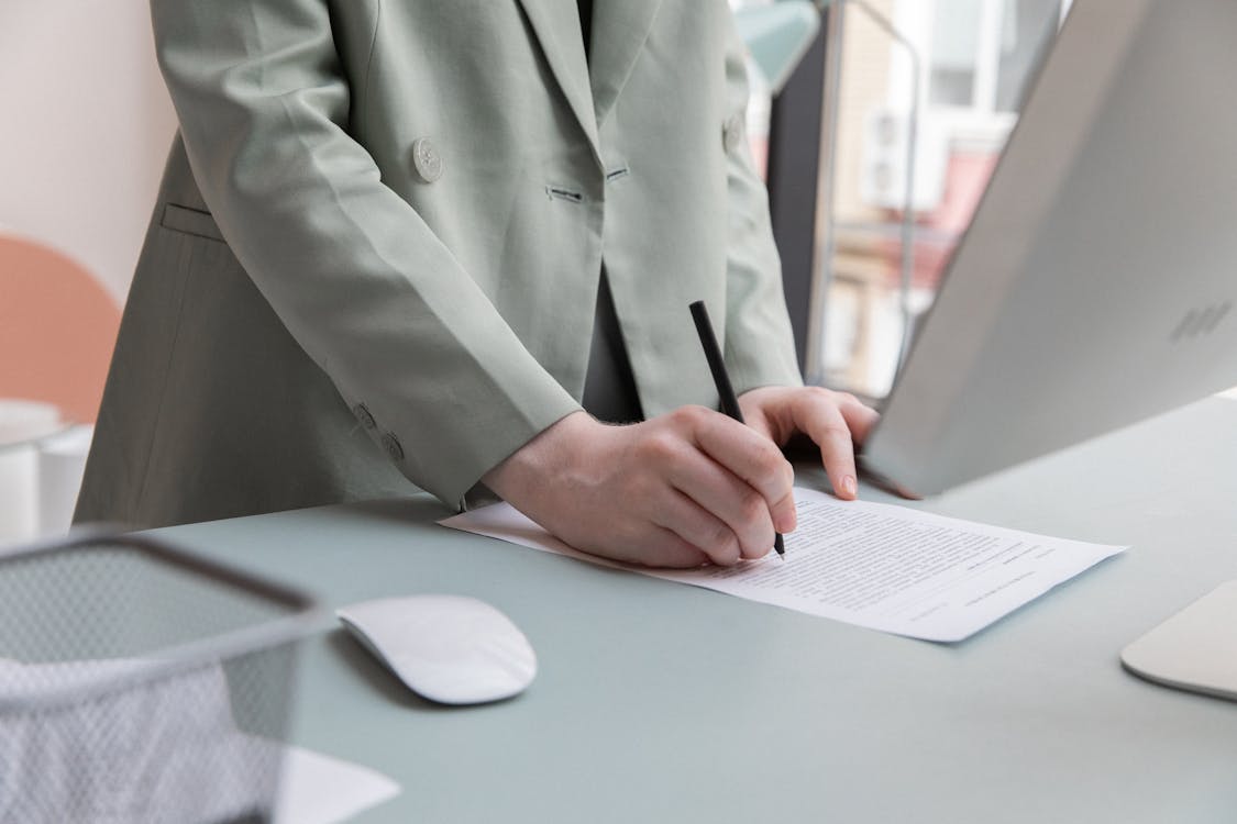 Free Unrecognizable worker standing at table with computer while taking notes in document while working in office Stock Photo