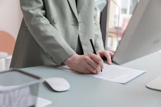 Unrecognizable worker standing at table with computer while taking notes in document while working in office