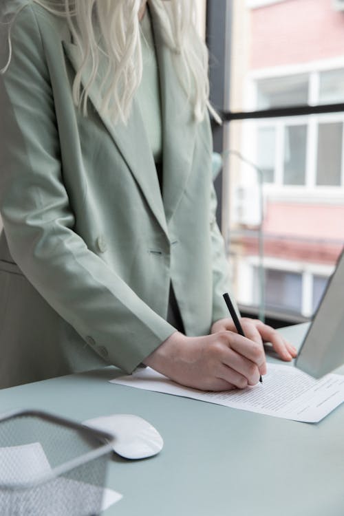 Free Crop faceless woman in formal clothes holding pen and writing notes on paper while working at table with computer and mouse Stock Photo