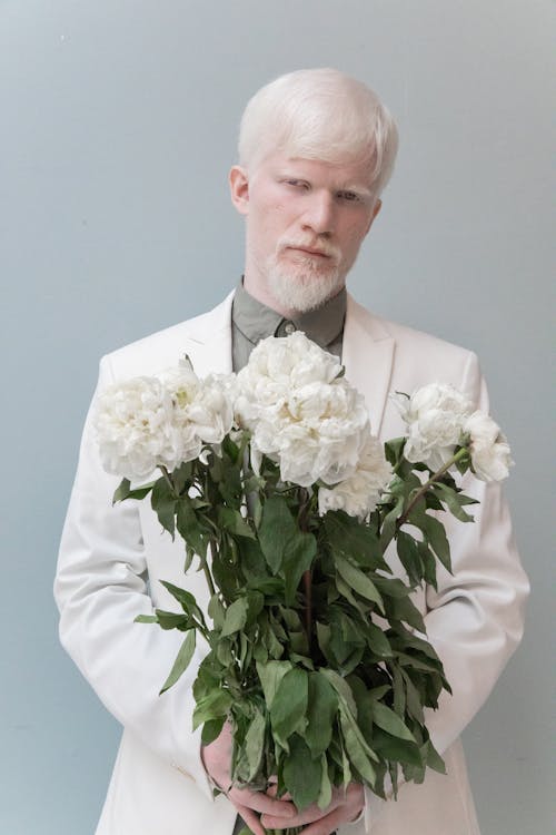 Confident blond male in formal suit holding bouquet of fresh flowers while standing against gray background and looking at camera