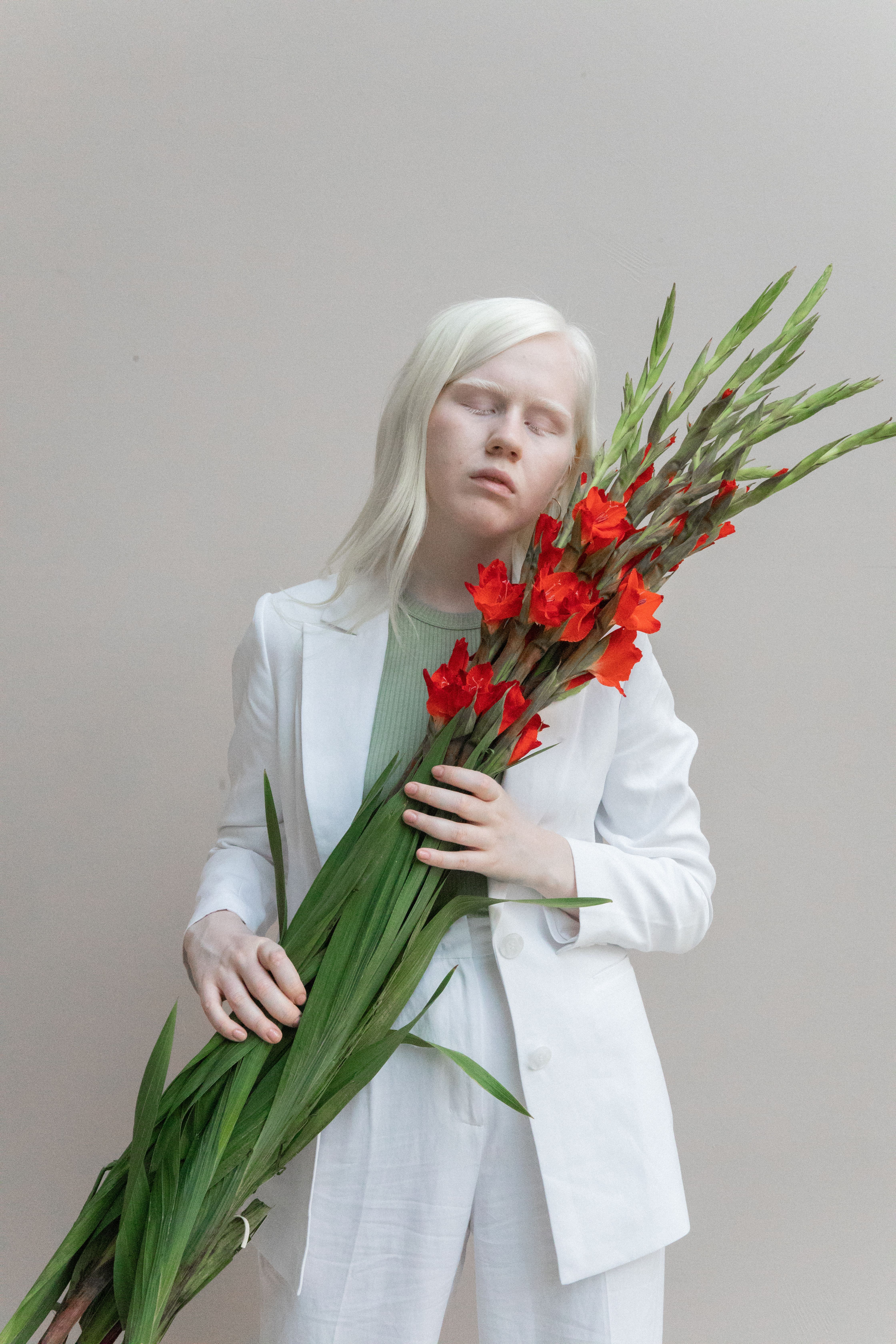 Woman in a white suit with closed eyes, holding vibrant red flowers.
