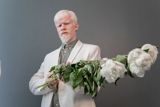 Low angle of serious blond haired male in white suit holding bouquet of blooming flowers while standing against gray background and looking at camera