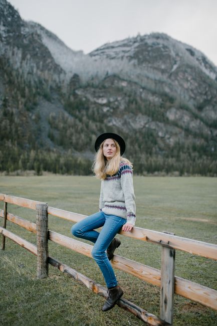 Full body of young female in casual outfit and hat leaning on wooden fence in mountainous terrain